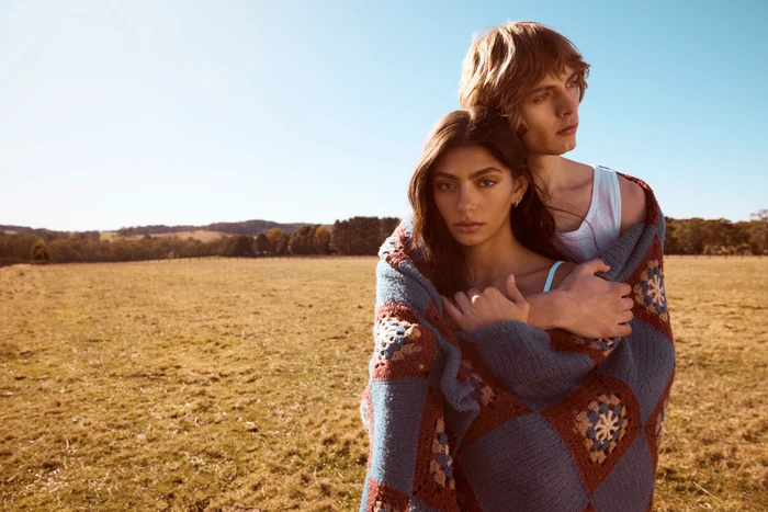 Two models wrapped in a blue and brown crocheted blanket standing in an open dry field under a warm sun