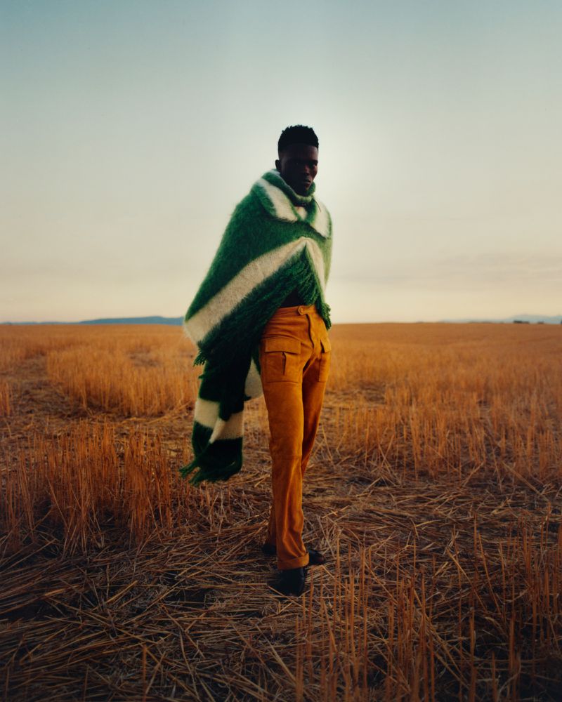 Person wrapped in a green and white shawl stands in a golden field during daytime, wearing mustard-colored pants in outdoor setting.