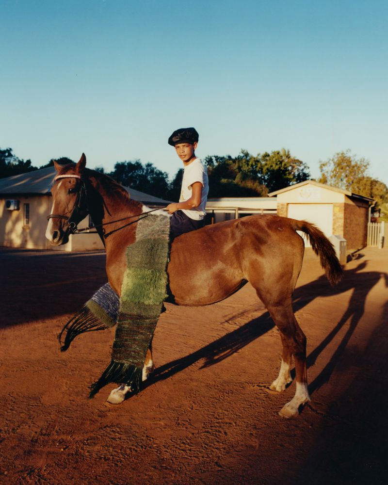 Young person sits on a horse outdoors, draped with a colorful woven shawl or blanket in an eye-level shot on a sunny day.