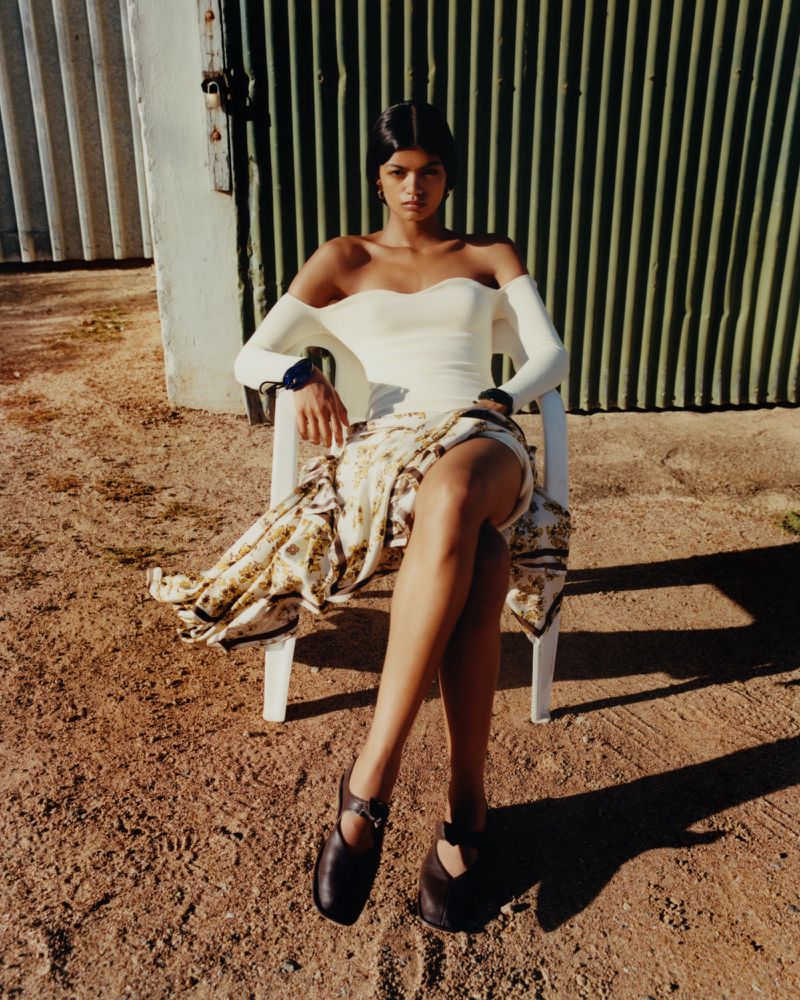 A tanned-skin female model poses sitting on a white chair in a cream-colored top, patterned wrap skirt, and dark flats outdoors.