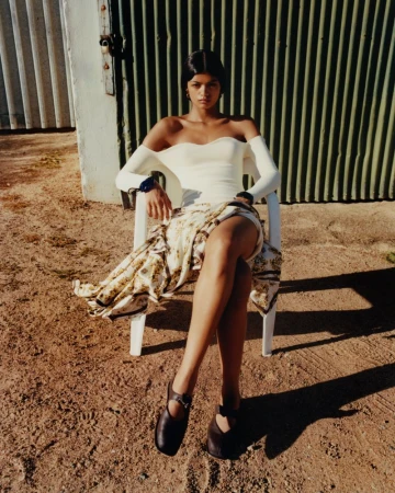 A tanned-skin female model poses sitting on a white chair in a cream-colored top, patterned wrap skirt, and dark flats outdoors.