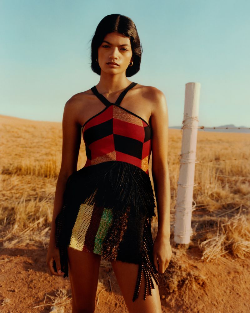 Model wearing a red and black geometric knit halter garment with black fringe detailing standing outdoors by a fence post.