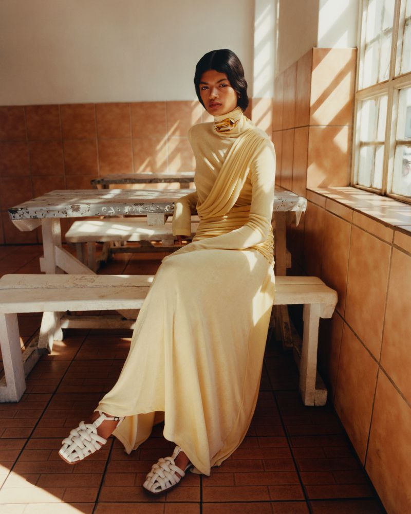 A young woman in a light yellow dress sits on a white bench in an indoor setting with natural light and tiled walls.