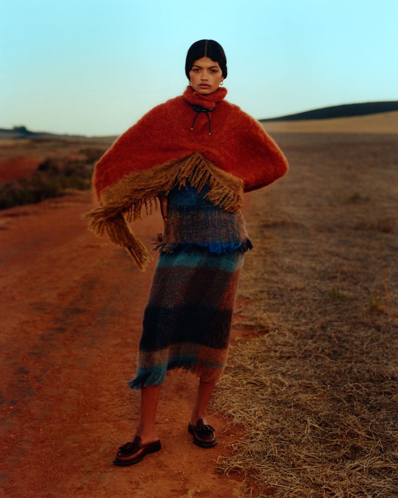 A stylish woman in an orange poncho and striped skirt stands in a field with a clear sky and a blurred horizon, showcasing fashion.
