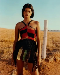 Model wearing a red and black geometric knit halter garment with black fringe detailing standing outdoors by a fence post.