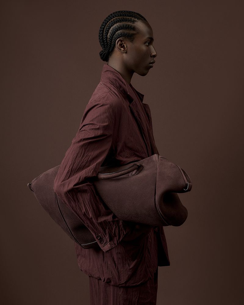 Fashion portrait of a person with braided hair, wearing a matching auburn jacket and pants, and carrying an oversized auburn bag.