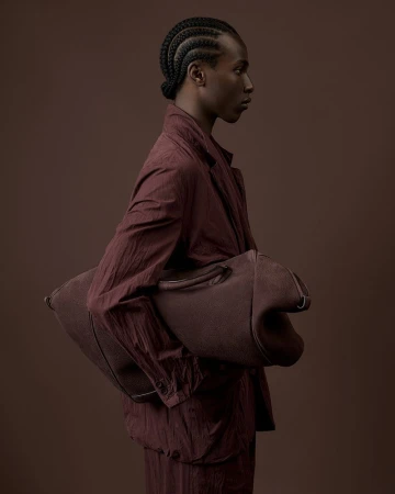 Fashion portrait of a person with braided hair, wearing a matching auburn jacket and pants, and carrying an oversized auburn bag.
