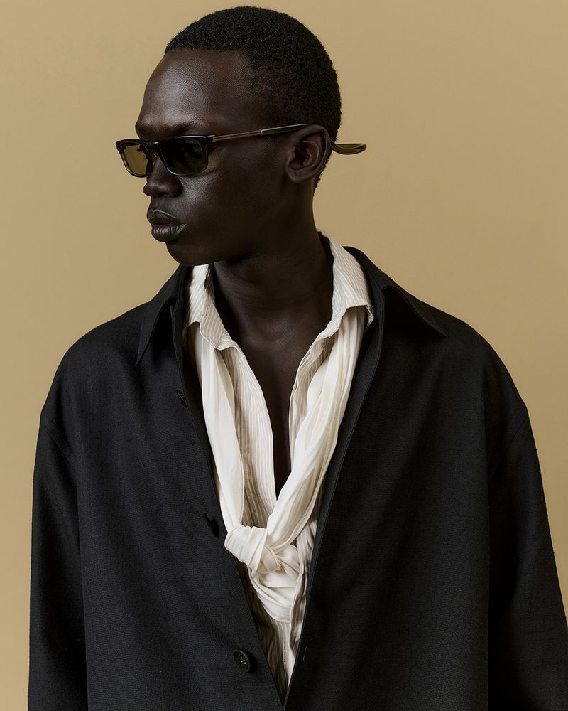 Dark-skinned male fashion model wearing sunglasses, black jacket, and draped collared shirt in a studio portrait against a tan backdrop.