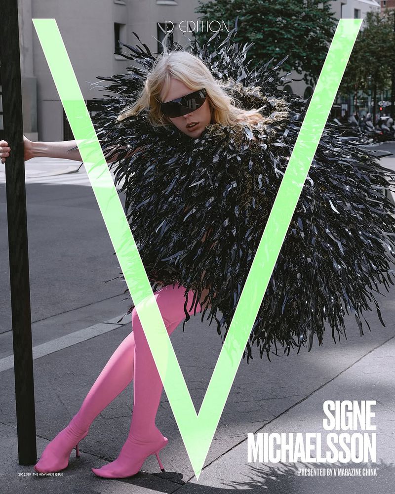 Editorial fashion shot of a woman in a feather coat and pink boots, posing against an urban street with building and large letter V.