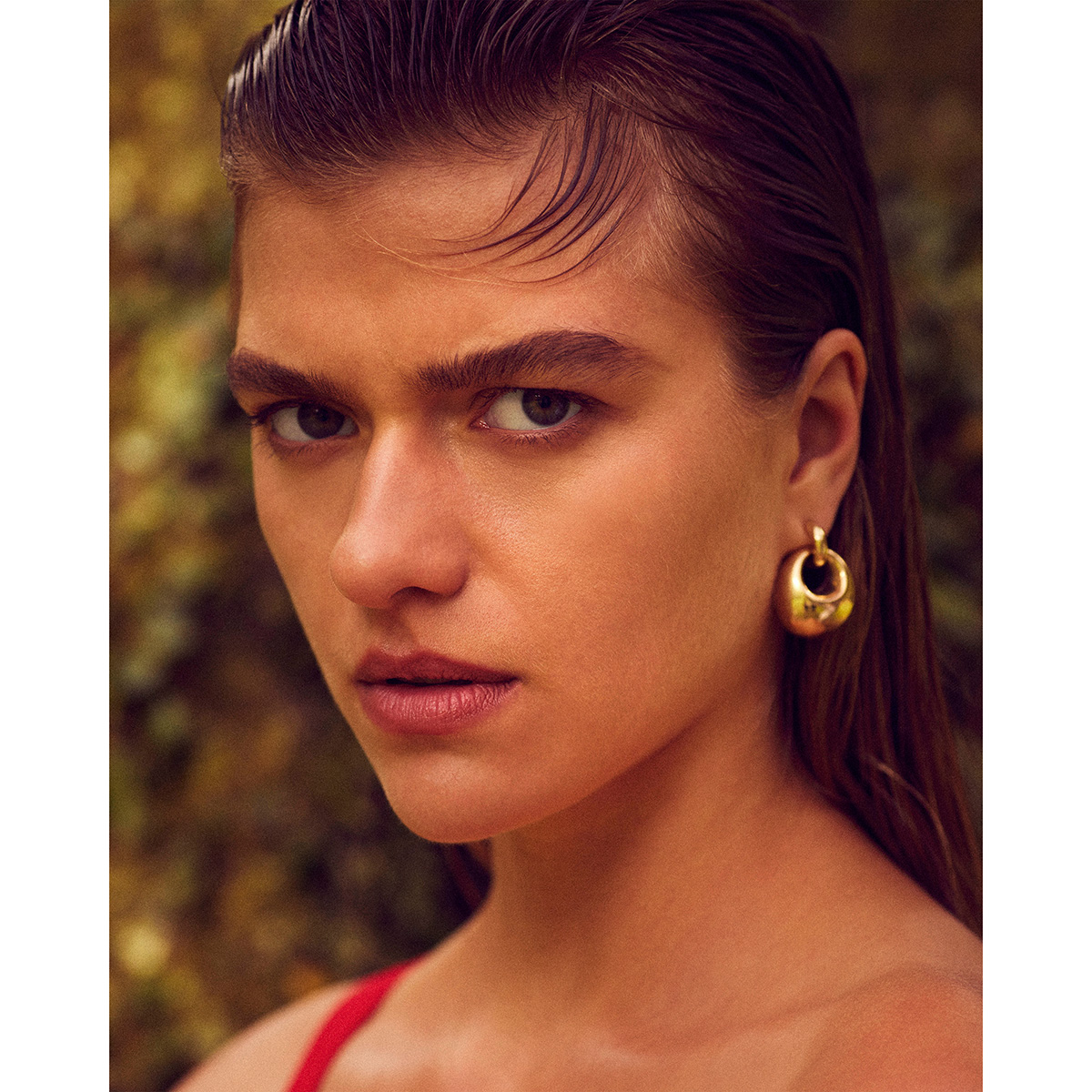 Close-up studio shot of a young woman with wet slicked-back hair, wearing gold hoop earrings and a red top against a blurred background.