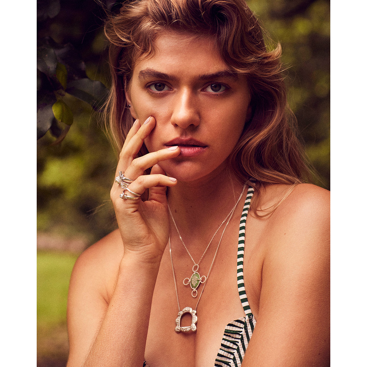 Close-up shot of a female model with long wavy hair, wearing rings and necklaces, posing outdoors in a natural, green environment.