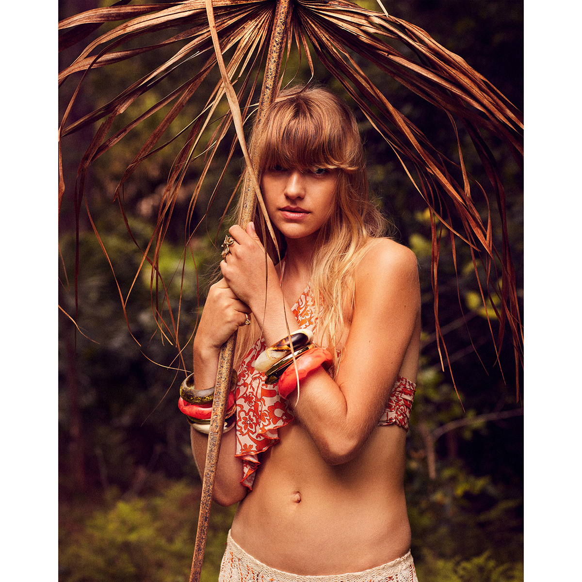 Young woman poses in bikini top holding a dried palm leaf in a lush outdoor environment, accessorized with multiple bracelets.