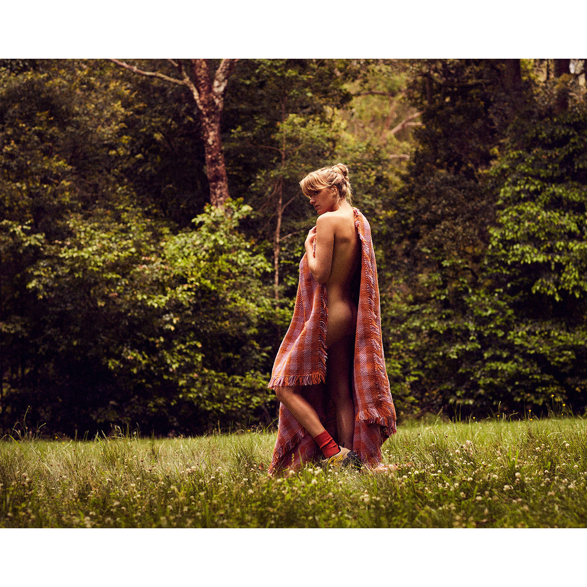 Woman draped in orange blanket posing in sunny field with green trees in the background.