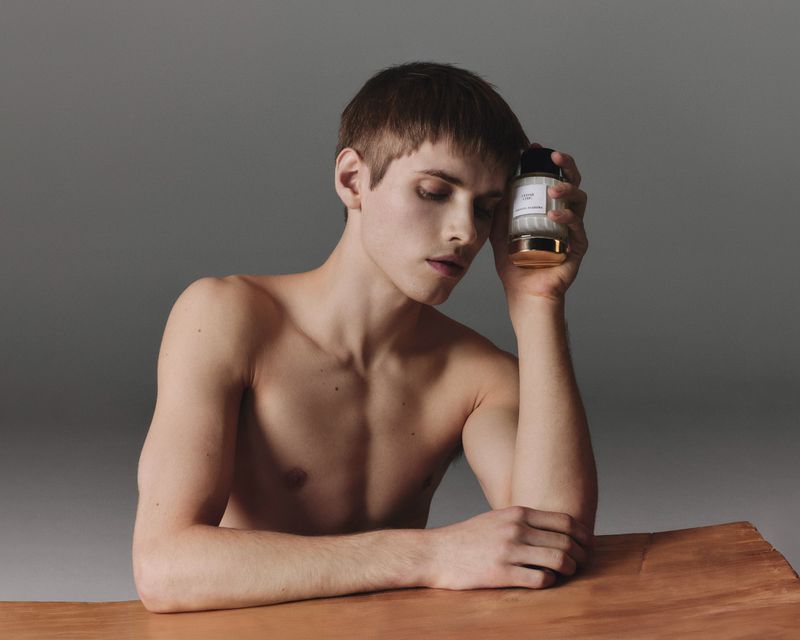 A bare-chested young adult male poses indoors holding a white and gold cosmetic bottle, resting his head on his hand on a copper surface.