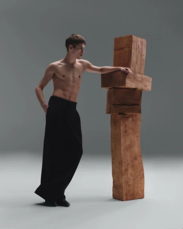 Shirtless male model in black pants poses next to a stacked wooden block structure against a gradient grey studio backdrop with soft lighting.