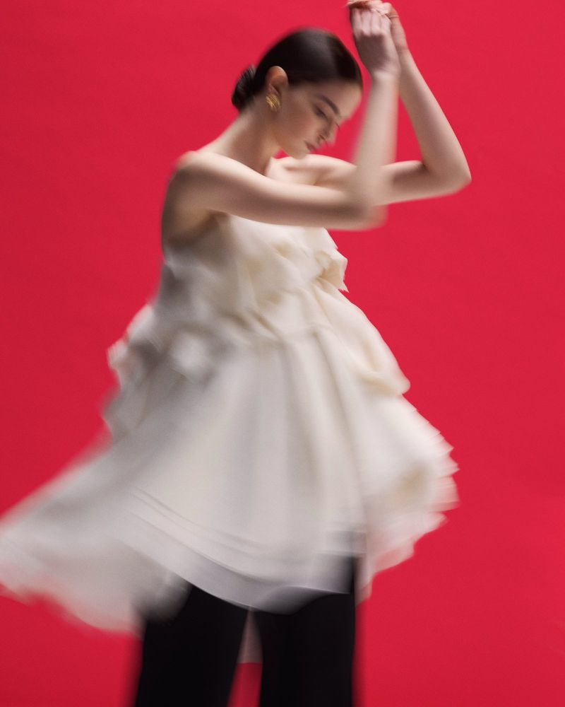 Woman in a cream-colored, ruffled dress and black pants, captured in motion against a vivid red background, creating a blurred effect.