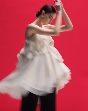 A model in motion wearing a voluminous tiered white strapless top and black trousers against a bold red studio background.