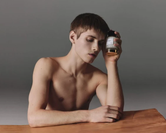 Shirtless male model leaning on a wooden table while holding a Carolina Herrera fragrance bottle against a grey studio backdrop.