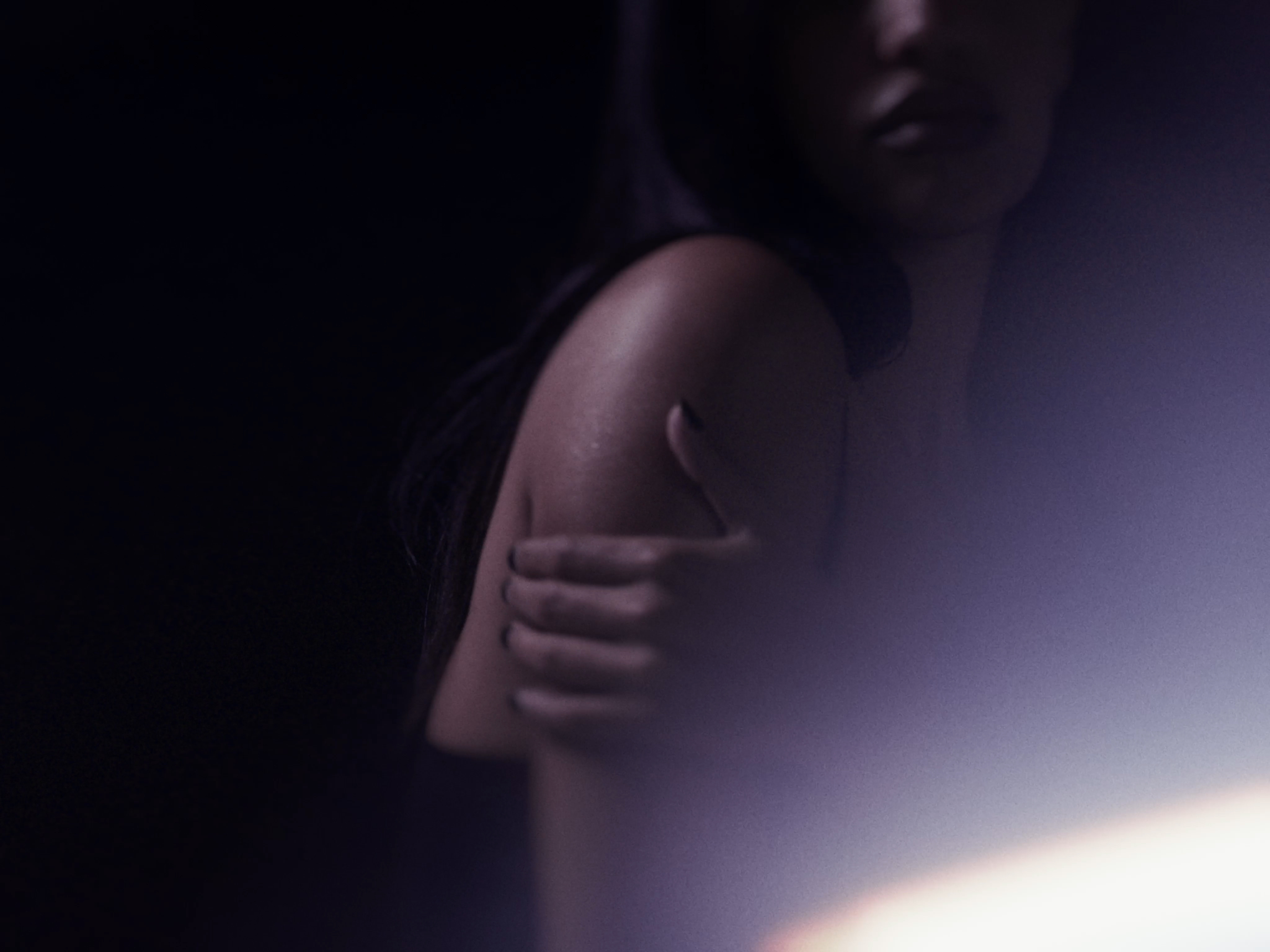 Cinematic low-key shot of a woman holding her bare arm with dark manicure in dim lighting and soft lens flare.