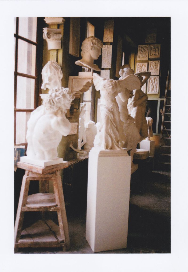 Photo studio interior filled with plaster sculptures, including busts on stands, against a backdrop of windows and architectural details.
