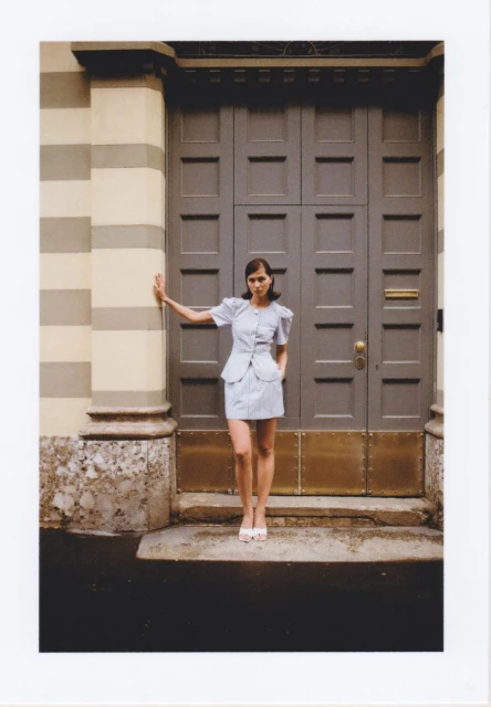A slender young woman in a light blue dress and white heels poses in front of a large grey paneled door and cream and grey striped wall.