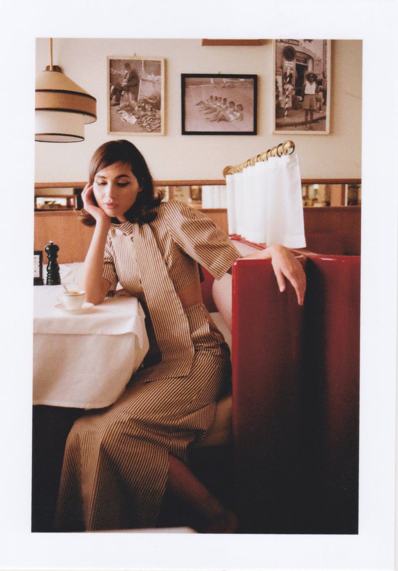 Young woman in striped dress sits in a red booth at a restaurant with black and white framed photos on the wall.
