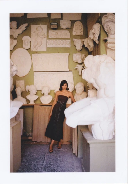 Fashion shot of a woman in a dark dress posing among white plaster sculptures in an indoor studio with neutral green walls.