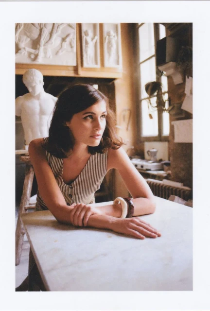 Woman in striped vest poses at marble table in artist's studio with sculptures, natural light, and neutral tones for fashion content.