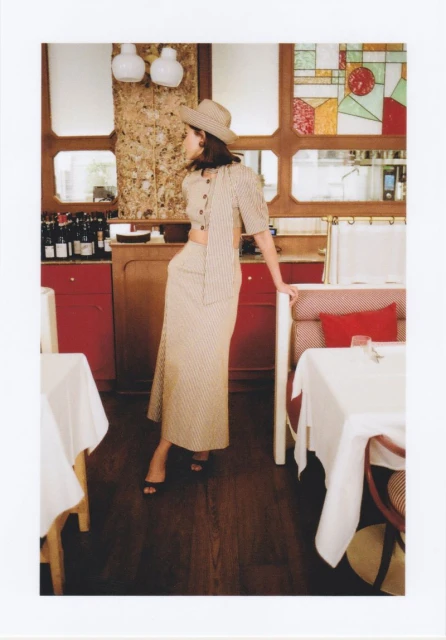 Woman in beige skirt set and hat in a restaurant setting with tables and chairs, red accents, decorative windows and wall details.