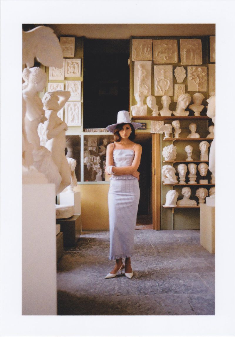 A slender woman poses in a fitted lavender dress and matching hat in an indoor art studio filled with plaster sculptures and busts.