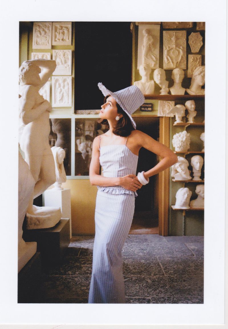 Woman in striped dress poses in front of art and sculptures at a museum, wearing a hat and bracelet, looking up with neutral expression.