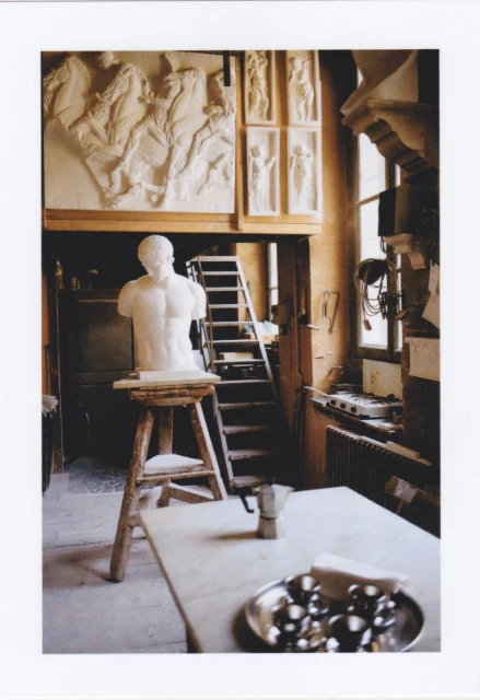 Sculpture studio with a white marble torso on a stand, surrounded by wall reliefs, tools, a table, and a ladder under natural light.