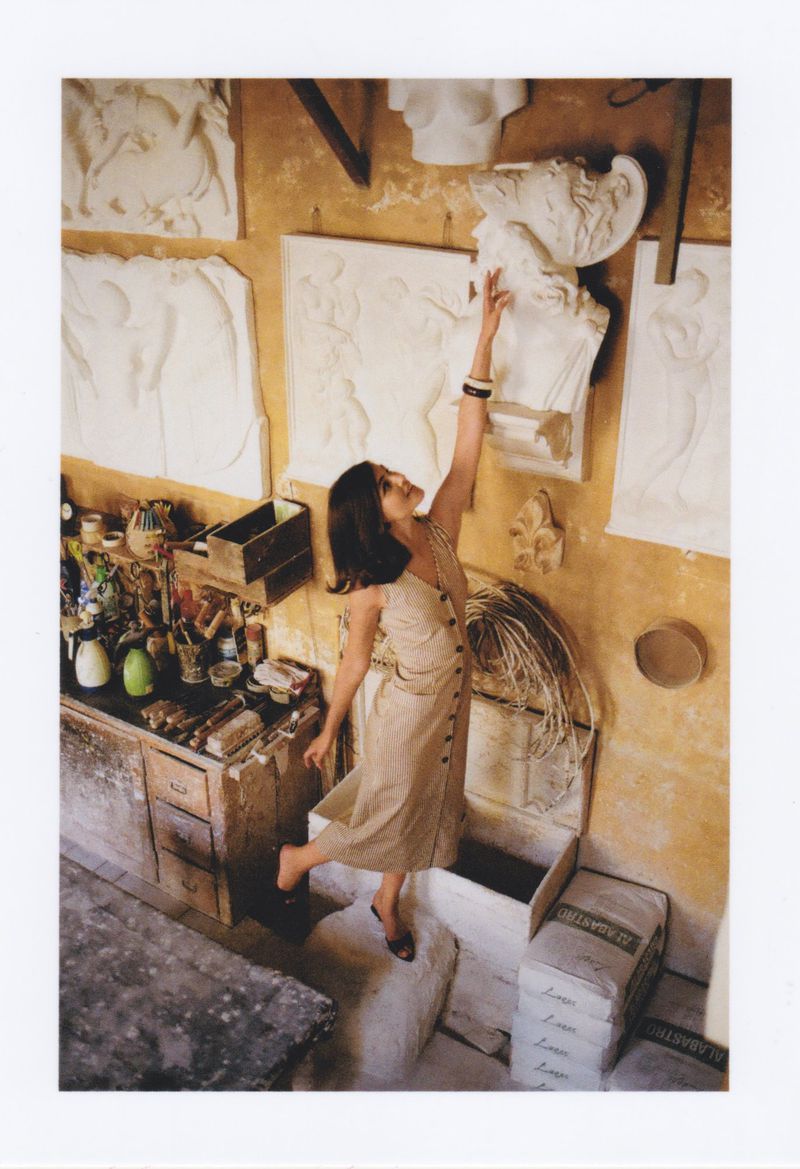Eye-level view of a brunette woman in a studio, reaching towards white bust sculptures on a tan wall, surrounded by art supplies.