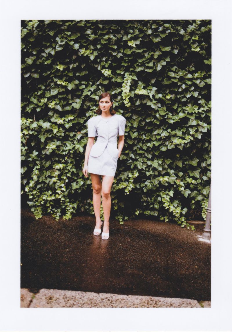 Full-length shot of an adult female fashion model posing outdoors in a light-blue mini-dress and white heels in front of a green ivy wall.
