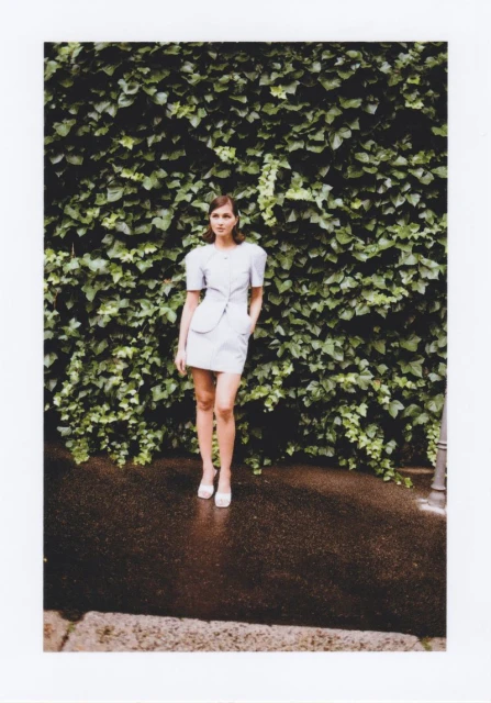 Full-length shot of an adult female fashion model posing outdoors in a light-blue mini-dress and white heels in front of a green ivy wall.