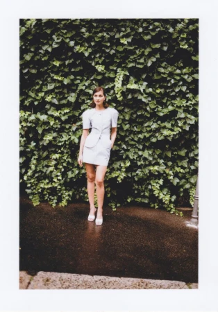 Full-length shot of an adult female fashion model posing outdoors in a light-blue mini-dress and white heels in front of a green ivy wall.