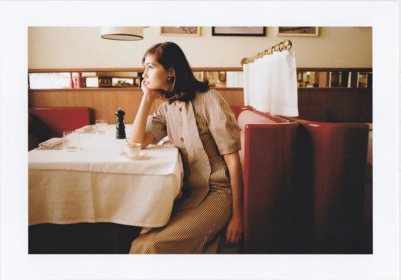 A woman sits in a red booth at a restaurant with a white tablecloth and a cup of coffee, looking away in a contemplative pose.