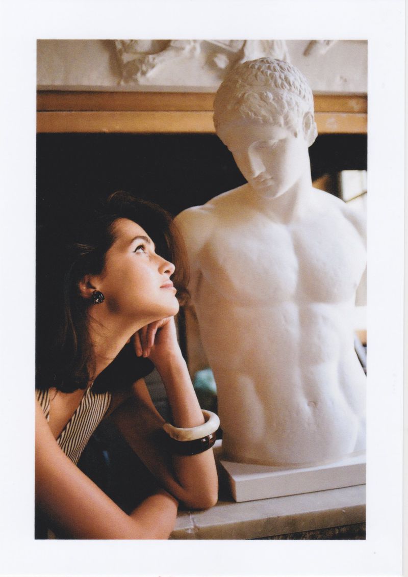 Elegant woman gazes at a white marble sculpture in an indoor setting, creating a harmonious blend of classic art and modern beauty.