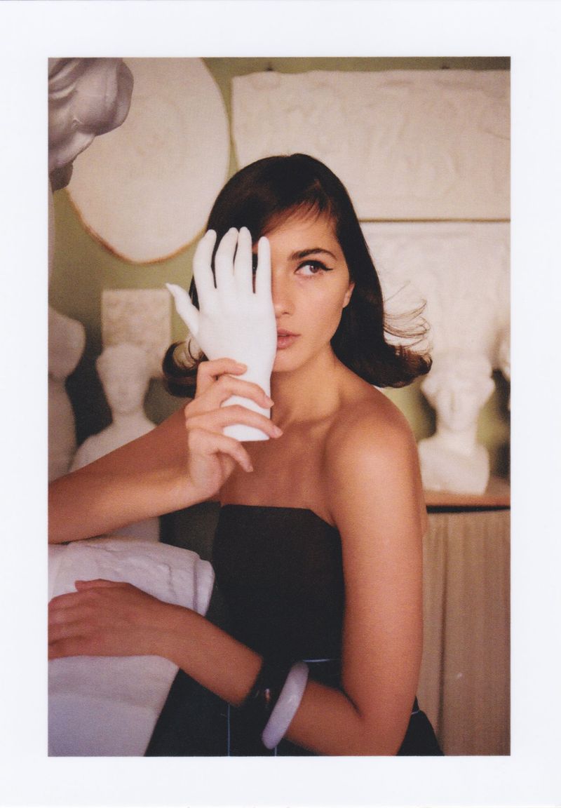 Brunette woman in a strapless black dress poses indoors holding a white glove up to her face, surrounded by white sculptures.