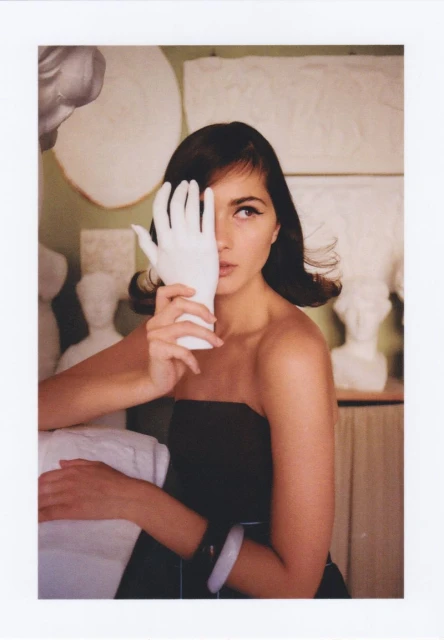 Brunette woman in a strapless black dress poses indoors holding a white glove up to her face, surrounded by white sculptures.