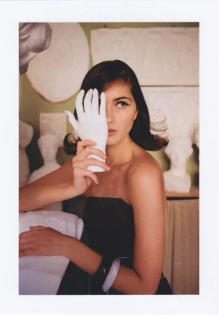 Brunette woman in a strapless black dress poses indoors holding a white glove up to her face, surrounded by white sculptures.