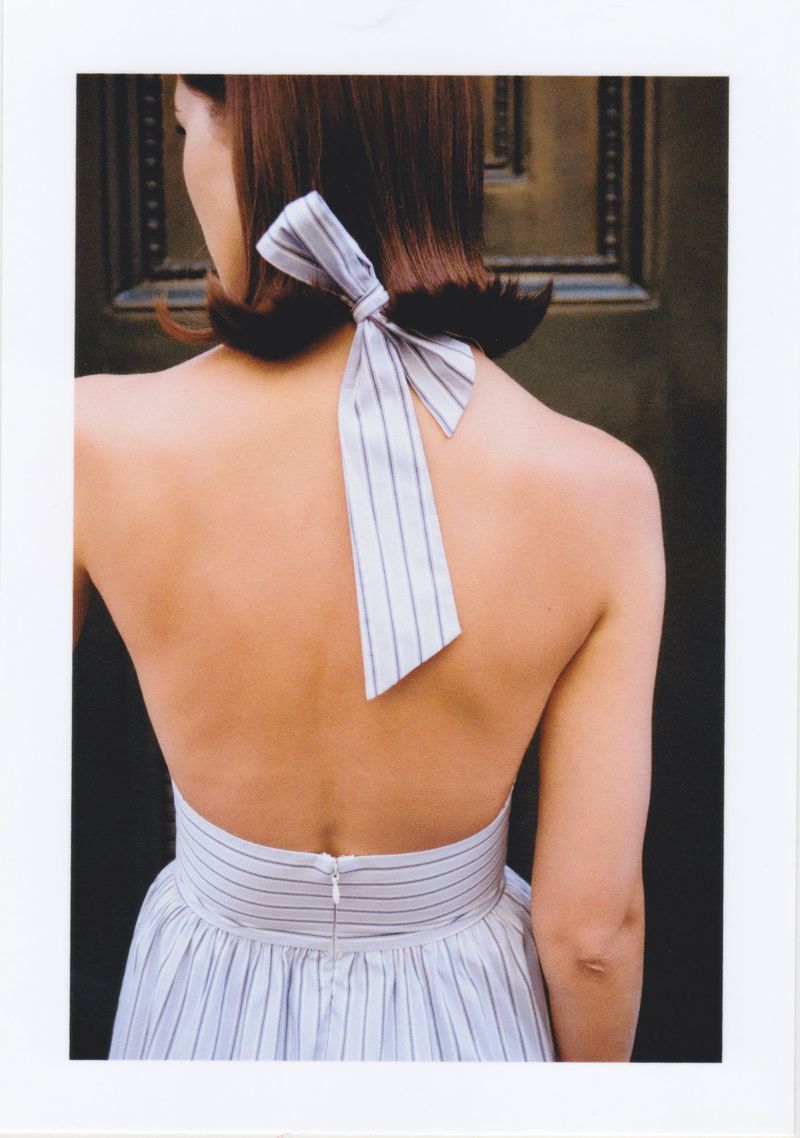 Back view of an adult female in a striped halter dress with a large bow at the nape, set against a dark paneled background.