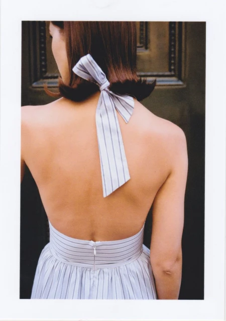 Back view of an adult female in a striped halter dress with a large bow at the nape, set against a dark paneled background.