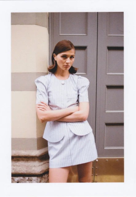 Stylish woman in a pinstripe skirt suit with arms crossed, leaning against a wall with a doorway in the background, fashion editorial.