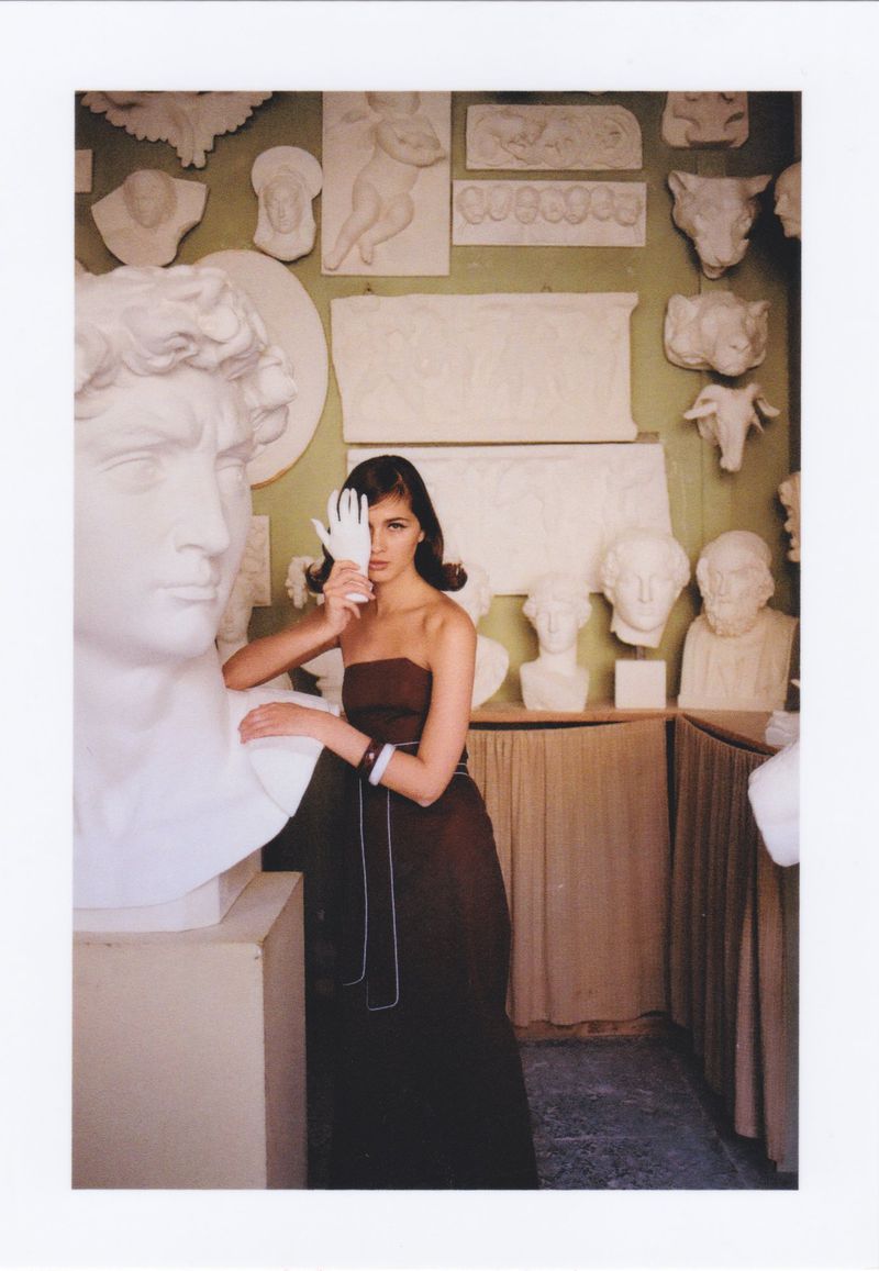 Fashion shot of a woman in a strapless gown, with a white glove over her eye, leaning on a white sculpture bust in an art gallery.