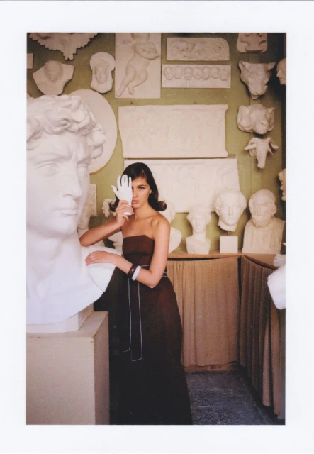 Fashion shot of a woman in a strapless gown, with a white glove over her eye, leaning on a white sculpture bust in an art gallery.