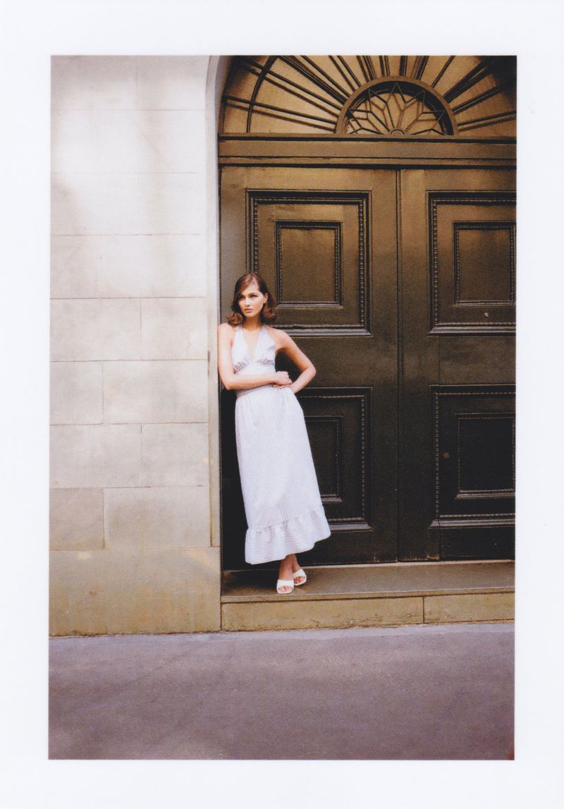 Elegant adult female model in white dress poses in front of grand, dark door with stone architrave and wall.