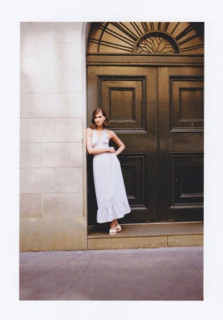 Elegant adult female model in white dress poses in front of grand, dark door with stone architrave and wall.