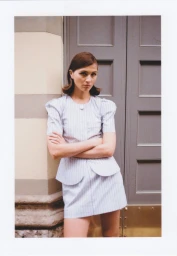 Model with retro flipped hair wearing a pale pinstripe short-sleeve mini skirt suit with arms crossed in front of a grey door.