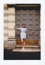 Woman in a light blue puff-sleeve skirt suit and white heels poses in front of a tall grey paneled doorway.